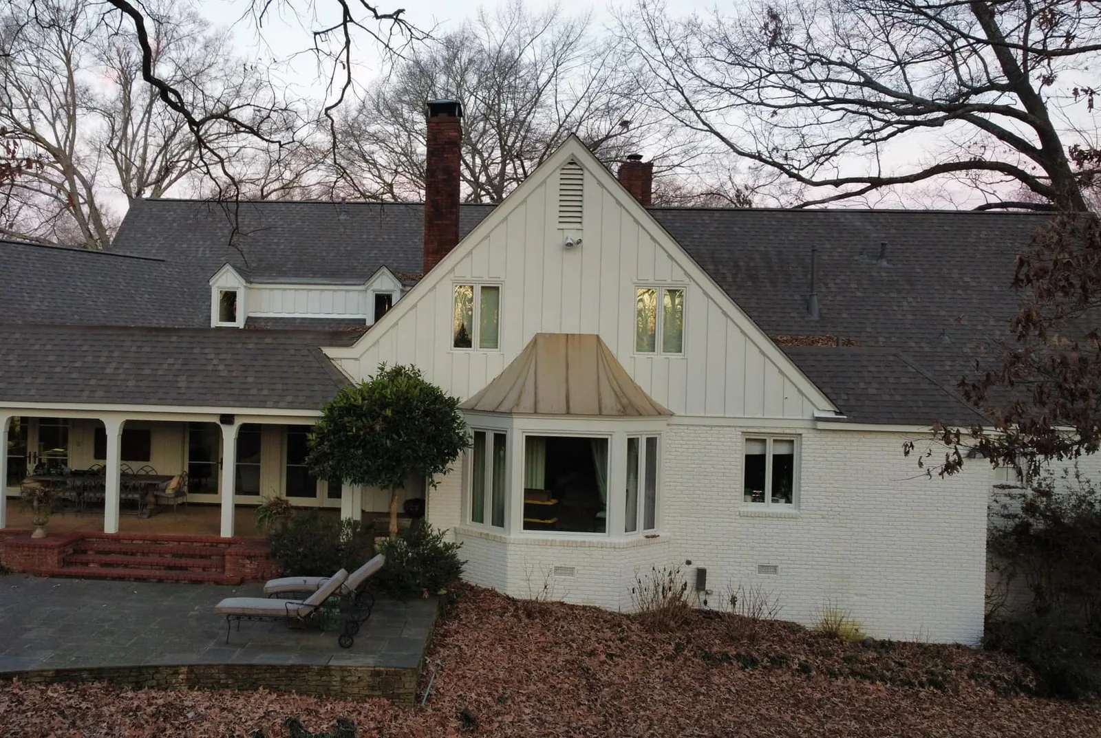 Back of a white painted brick and board-and-batten home with fallen leaves and bare trees in late fall